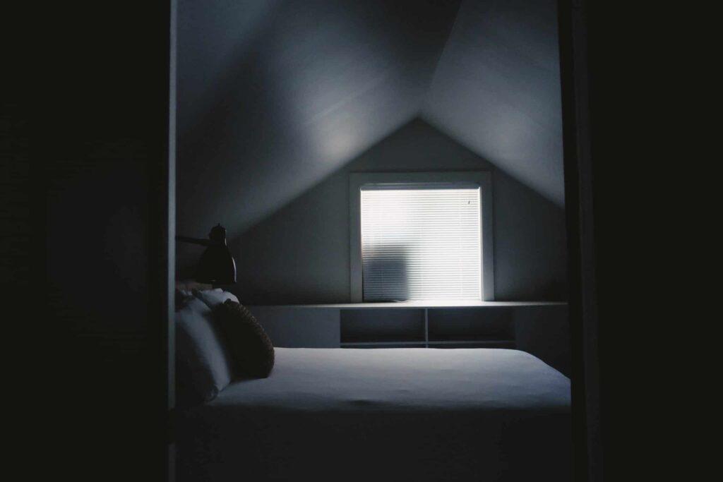 A cozy attic bedroom with a neatly made bed, soft lighting from a bedside lamp, and gentle daylight streaming through closed blinds. At Bell Brothers, our trusted background-checked technicians are dedicated to keeping every room in your home as comfortable as this one—backed by our No Surprise Guarantee and clear, upfront pricing. Proudly serving Northern California communities with reliable service you can count on.