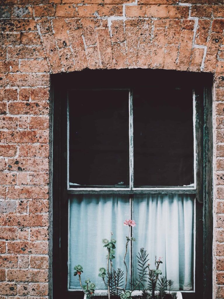 A charming vintage brick wall features a wooden-framed window with a white curtain, surrounded by tall green plants. Just like this inviting scene, Bell Brothers brings reliability and comfort to homes across Northern California. With our No Surprise Guarantee, background-checked technicians, and transparent upfront pricing, you can trust us to deliver exceptional service every time—something our local communities have relied on for years.