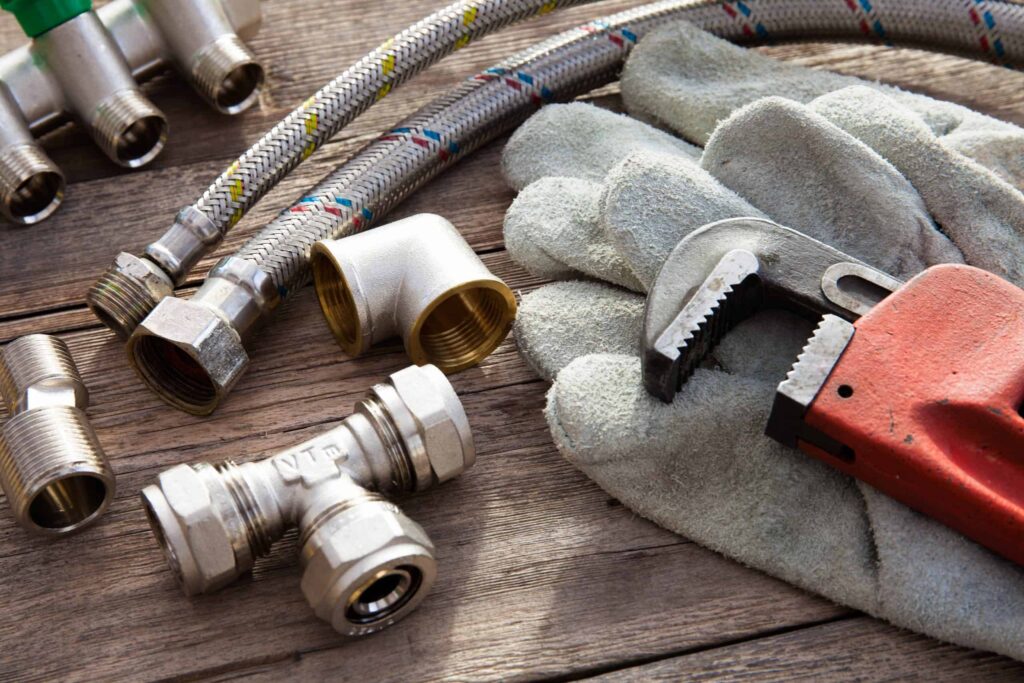 A selection of high-quality plumbing tools and fittings—including a pipe wrench, flexible hoses, metal connectors, and protective work gloves—neatly arranged on a wooden surface. Bell Brothers’ trusted, background-checked technicians use these professional tools to deliver expert service to Northern California communities. With our No Surprise Guarantee and upfront pricing, you can count on honest work and peace of mind every time.