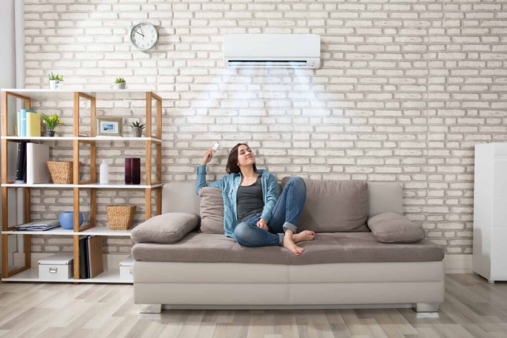 A woman relaxes on a comfortable gray couch, using a remote to easily control her wall-mounted air conditioner as cool air fills her modern living room. With Bell Brothers’ No Surprise Guarantee, upfront pricing, and background-checked technicians, homeowners across Northern California trust us to keep their homes comfortable—all with no unexpected costs or hassles.