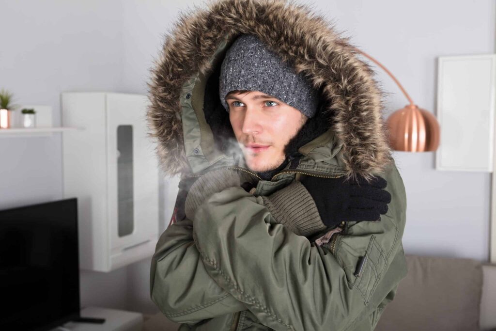 A person bundled up in a winter coat, fur-lined hood, knit hat, and gloves looks chilly indoors—breath visible in the cold air. With Bell Brothers’ background-checked technicians and No Surprise Guarantee offering upfront pricing, families across Northern California trust us to keep their homes cozy all season long.