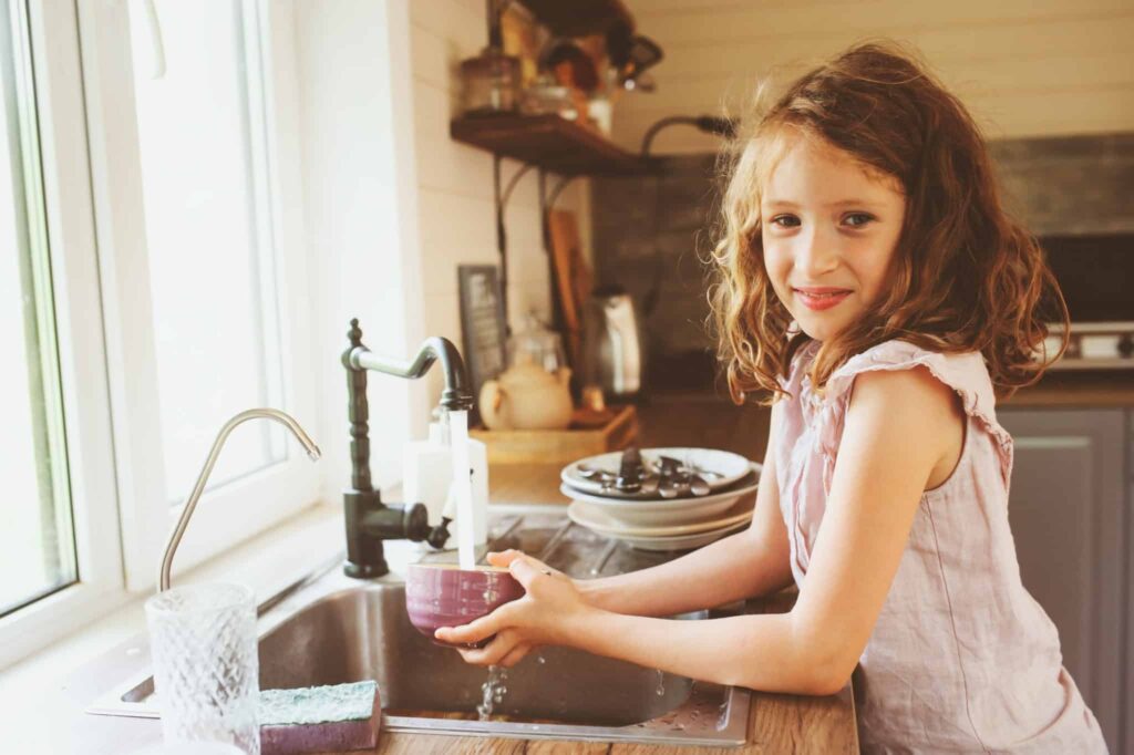 A young girl washes a purple bowl at the kitchen sink, surrounded by neatly stacked dishes and a sponge. At Bell Brothers, our trusted, background-checked technicians bring this same care to your home, proudly serving Northern California communities with upfront pricing and our No Surprise Guarantee.
