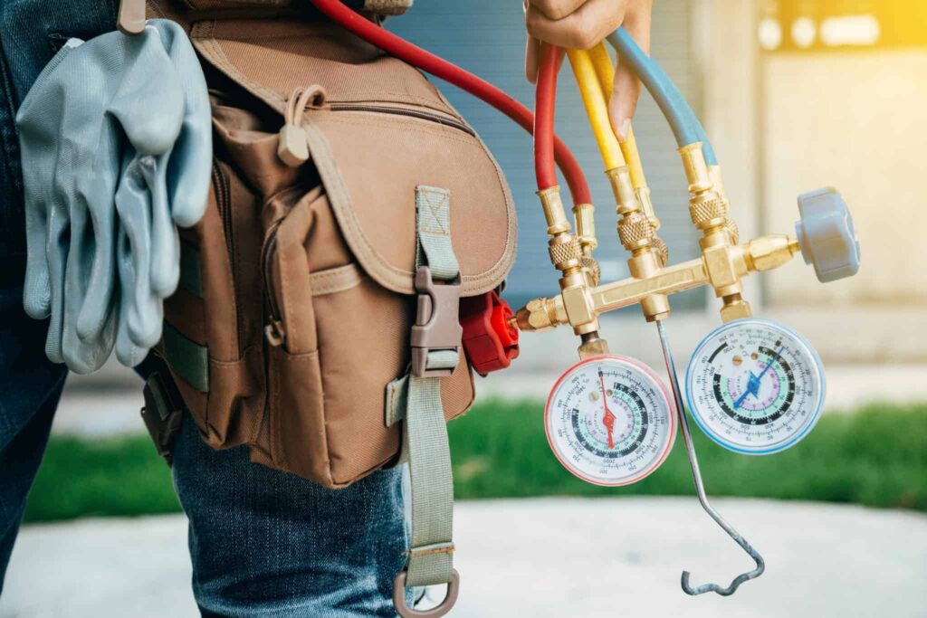 A Bell Brothers technician, fully background-checked and equipped with a professional tool bag and gloves, prepares for HVAC maintenance using a manifold gauge set. We proudly serve Northern California communities with our trusted No Surprise Guarantee, upfront pricing, and a reputation built on reliable service you can count on.
