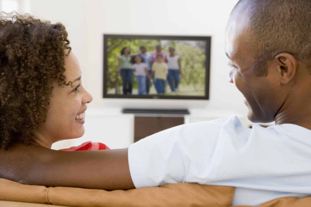 A smiling couple relaxes together on their living room couch, confident in their choice of Bell Brothers. The television behind them shows a happy family enjoying the outdoors—a reflection of the comfort and peace of mind Bell Brothers provides with background-checked technicians, upfront pricing, and a No Surprise Guarantee. Trusted throughout Northern California communities, Bell Brothers puts your family’s comfort first.