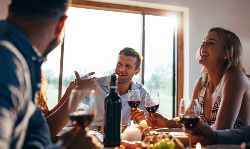 Four friends enjoying a meal together at a sunlit dining table, sharing smiles and conversation over red wine. Just like this gathering, Bell Brothers brings warmth and trust to Northern California homes with our No Surprise Guarantee, background-checked technicians, transparent upfront pricing, and a reputation for reliable service in your community.
