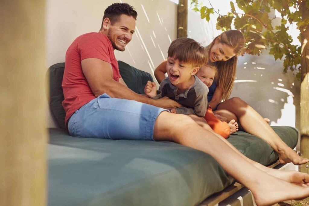 A happy family of four relaxes together on a cushioned outdoor bench, sharing smiles and laughter beneath the shade of leafy vines. Their comfort reflects the trusted service Bell Brothers provides throughout Northern California, with background-checked technicians, upfront pricing, and our No Surprise Guarantee—giving your family peace of mind every step of the way.