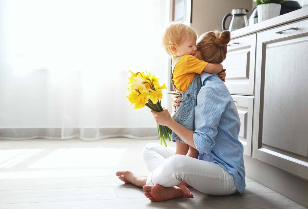 A smiling woman sits on her kitchen floor, embracing a young child and holding a cheerful bouquet of yellow flowers as sunlight streams through the window. With Bell Brothers, families across Northern California can enjoy moments like these with peace of mind, knowing our trusted technicians are background-checked and committed to delivering upfront pricing and our No Surprise Guarantee—just one reason we're a top choice in local communities.