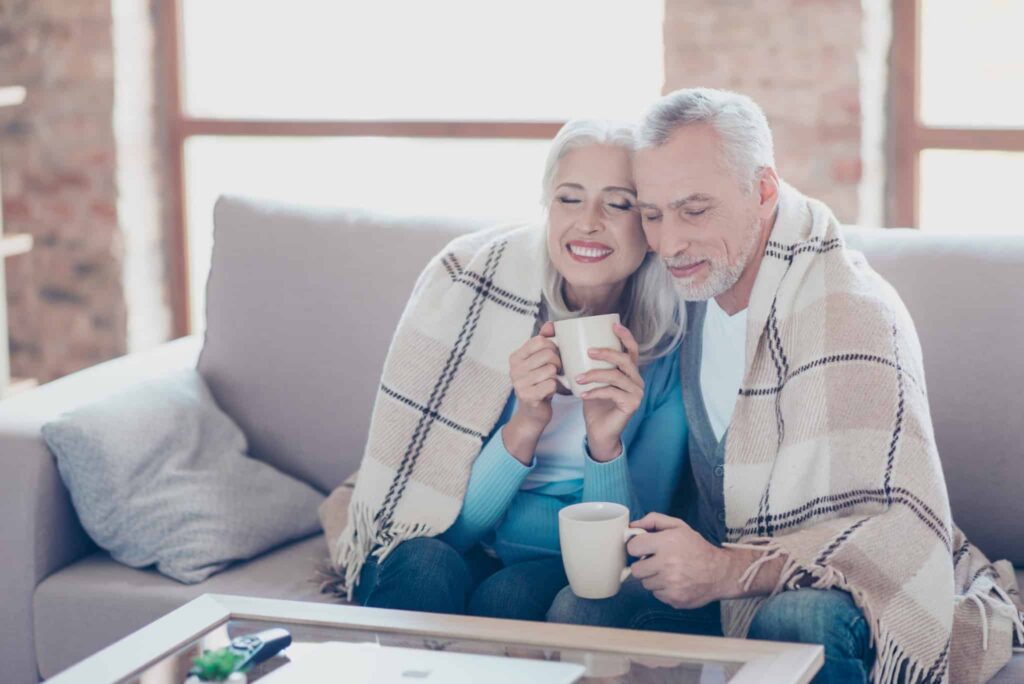 A smiling older couple relaxes together on their couch, wrapped in a cozy plaid blanket and enjoying hot drinks, confident in the comfort their home provides. The Bell Brothers No Surprise Guarantee ensures peace of mind with only background-checked technicians and clear, upfront pricing—no hidden fees. Trusted throughout Northern California communities for reliable service, Bell Brothers helps families just like theirs feel comfortable all year long.