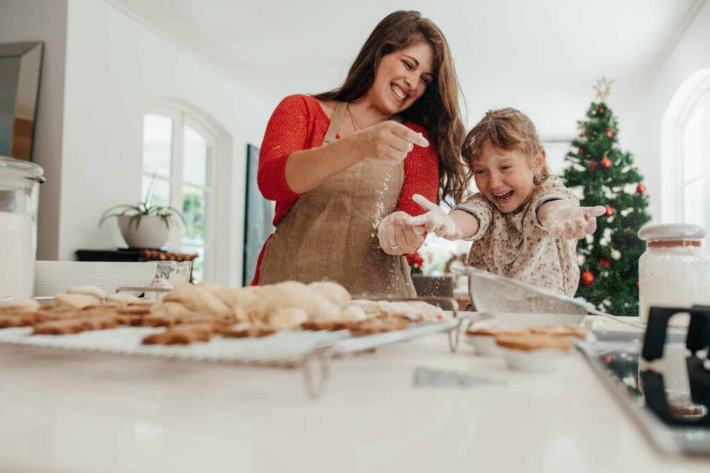 A smiling woman and a young girl wearing aprons bake cookies together in a bright, festive kitchen, playfully dusting each other with flour while a decorated Christmas tree glows in the background. Throughout Northern California, families like theirs trust Bell Brothers for reliable home comfort, thanks to our No Surprise Guarantee, fully background-checked technicians, upfront pricing, and strong reputation for friendly service.