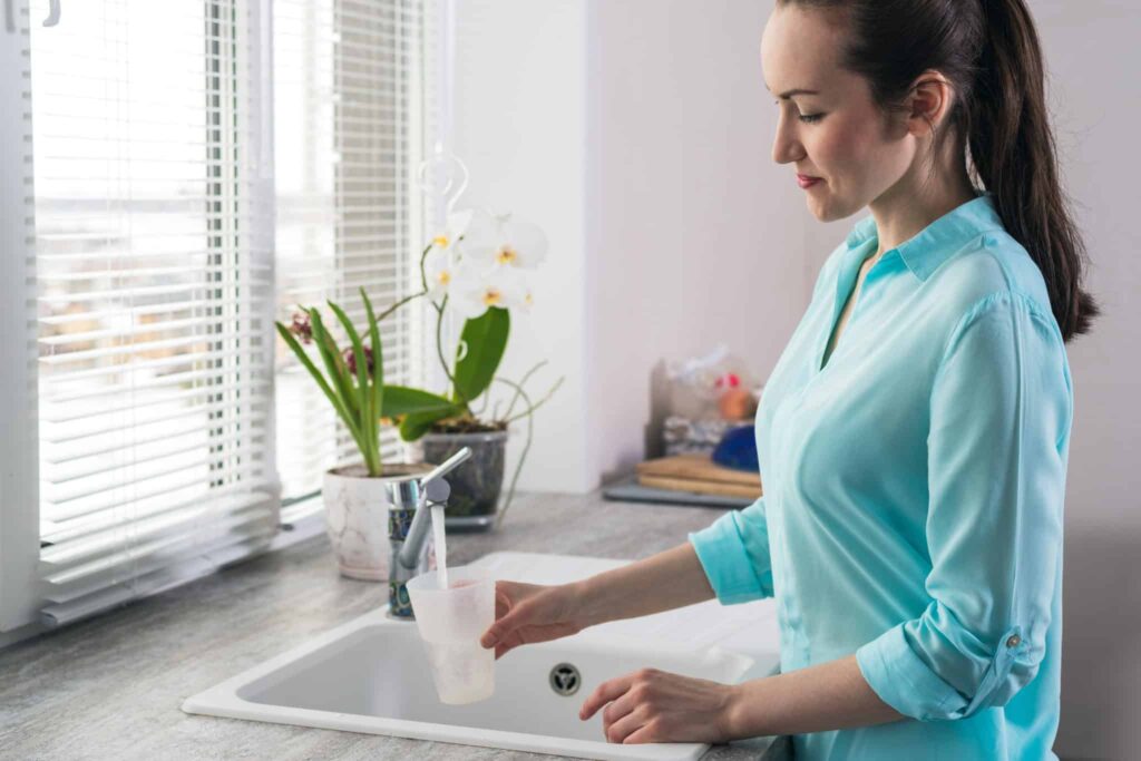 A woman in a blue shirt fills a cup with fresh water at her kitchen sink, surrounded by potted plants and natural light. Bell Brothers’ trusted, background-checked technicians provide expert service you can count on—backed by our No Surprise Guarantee, honest upfront pricing, and strong reputation for reliability across Northern California communities.