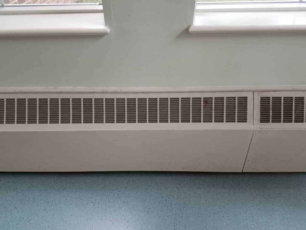 A clean, white wall-mounted radiator vent sits below two windows, set against a blue-speckled floor. At Bell Brothers, our trusted technicians—background-checked for your peace of mind—deliver friendly, professional service throughout Northern California. With upfront pricing and our No Surprise Guarantee, you can expect honest solutions and a comfortable home every time.