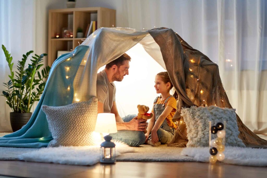 A parent and child relax together inside a cozy blanket fort decorated with glowing string lights, pillows, and a favorite stuffed toy—enjoying comfort and peace of mind thanks to Bell Brothers' trusted service. Proudly serving Northern California communities, Bell Brothers is known for background-checked technicians, upfront pricing, and our No Surprise Guarantee—so you can create warm moments at home with total confidence.
