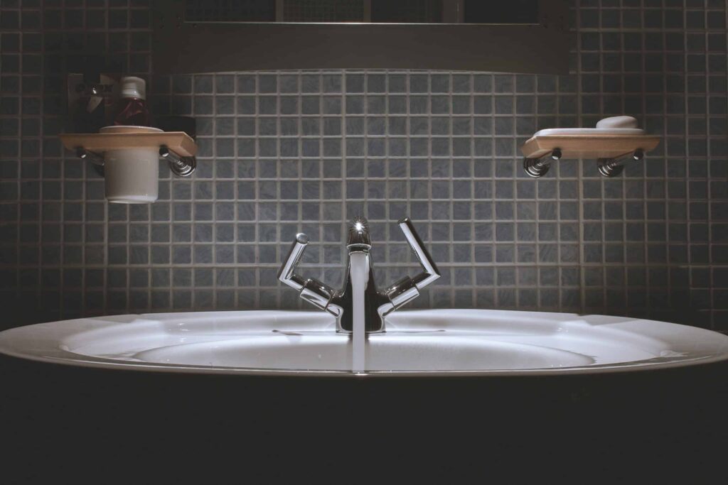A modern bathroom sink with a sleek chrome faucet is set against a blue tile backsplash, flanked by two wooden shelves stocked with toiletries. Trust Bell Brothers’ No Surprise Guarantee, background-checked technicians, and upfront pricing for reliable plumbing solutions throughout Northern California’s communities.