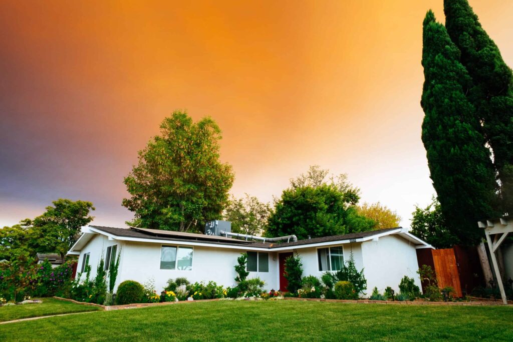 A beautifully maintained single-story white home with a lush lawn and garden is nestled among trees and shrubs beneath a glowing orange sunset. This scene reflects the trusted service Bell Brothers brings to Northern California communities — always with background-checked technicians, upfront pricing, and our signature No Surprise Guarantee for your complete peace of mind.