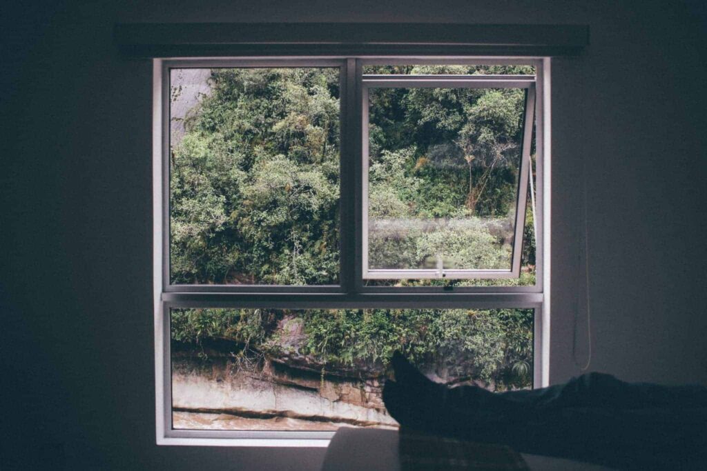 A bright window with one pane open reveals lush green trees and a rocky ledge outside. Inside, a person's legs and feet rest comfortably, suggesting peace of mind. At Bell Brothers, we bring this same comfort to your home with our trusted service—backed by our No Surprise Guarantee, background-checked technicians, and straightforward upfront pricing. Proudly serving Northern California communities with an outstanding local reputation you can count on.