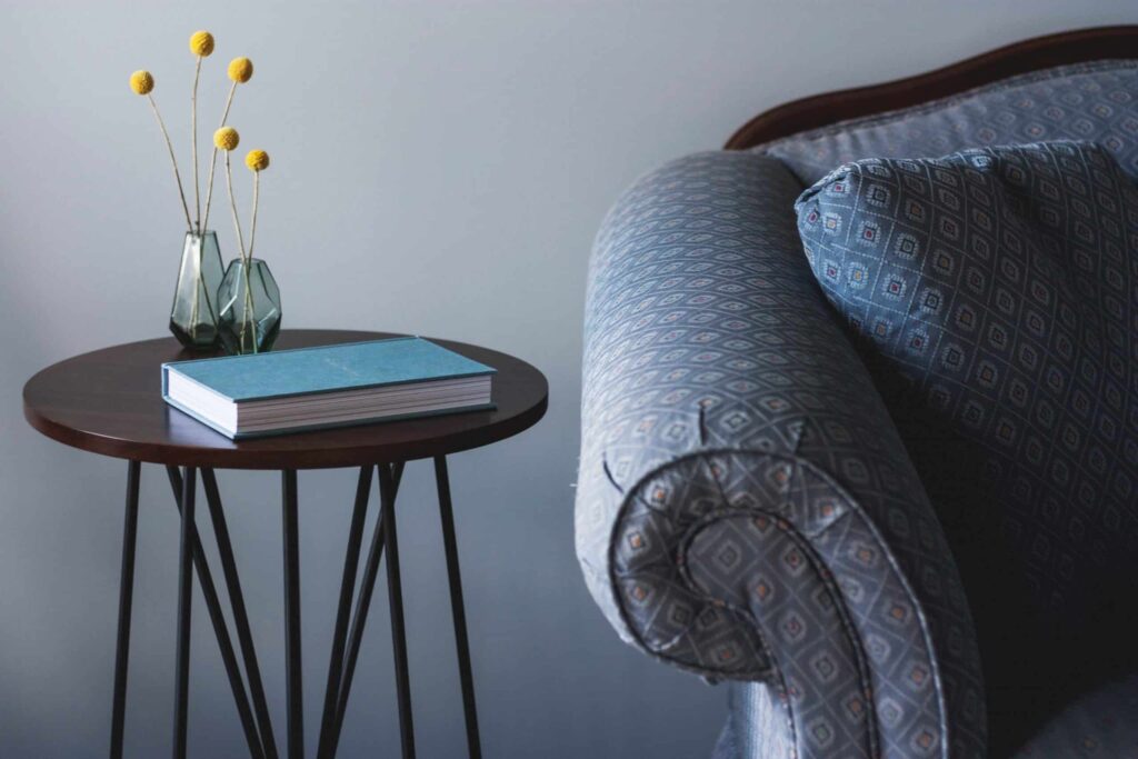 A round side table sits beside a blue patterned sofa, topped with a closed blue book and a cheerful vase of yellow billy buttons—reflecting the warmth and reliability Bell Brothers brings to homes across Northern California. Our trusted, background-checked technicians deliver upfront pricing and our No Surprise Guarantee to every community we serve.