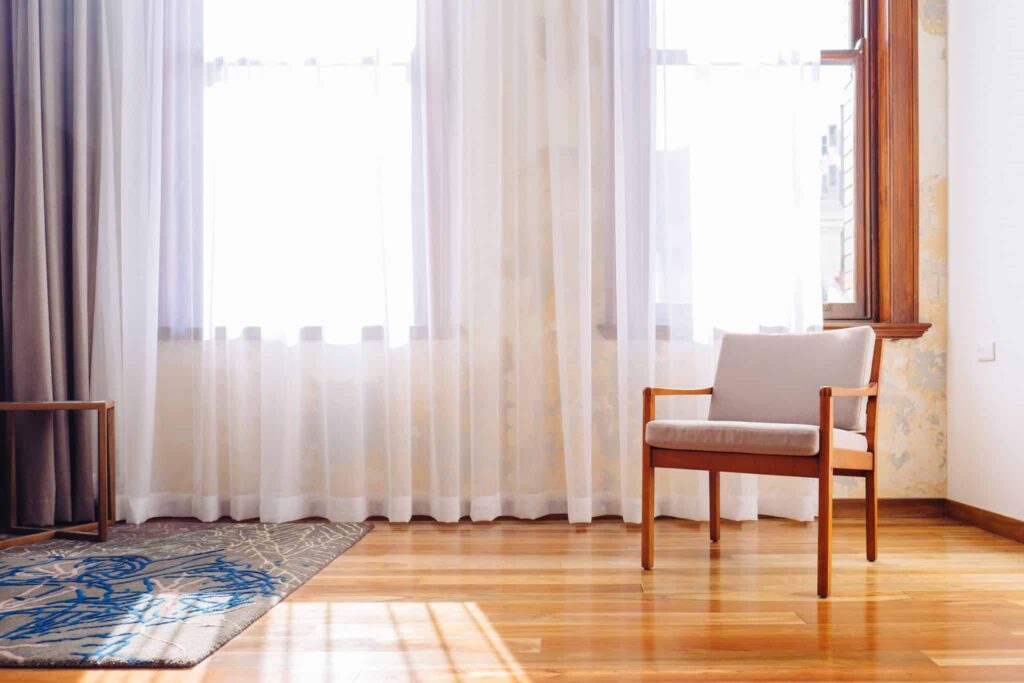 A sunlit room featuring a wooden chair with light cushions on a gleaming hardwood floor, illuminated by natural light through large windows with sheer white curtains. Just like this welcoming space, Bell Brothers brings comfort and reliability to your home—with our No Surprise Guarantee, background-checked technicians, clear upfront pricing, and a reputation for trusted service throughout Northern California communities.