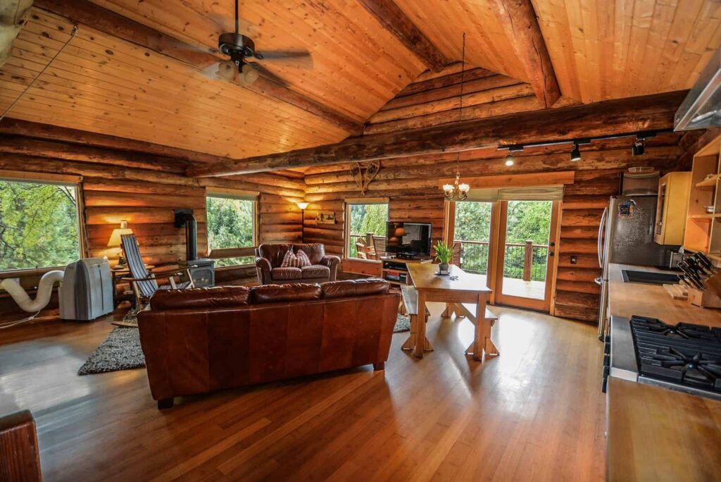 Spacious log cabin interior featuring gleaming wooden floors, comfortable leather sofas, and a modern kitchen area—all illuminated by large windows that showcase lush greenery outside. At Bell Brothers, our trusted technicians—each background-checked for your peace of mind—offer upfront pricing as part of our No Surprise Guarantee. Proudly serving Northern California communities with honest, reliable service you can count on.