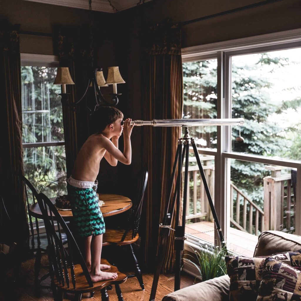 A young boy in swim trunks stands safely on a chair, gazing through a telescope out of a large window towards scenic trees and a sunlit deck. Just as he explores new horizons, families across Northern California trust Bell Brothers for worry-free home services—thanks to our No Surprise Guarantee, background-checked technicians, upfront pricing, and strong reputation for dependable care in every community we serve.