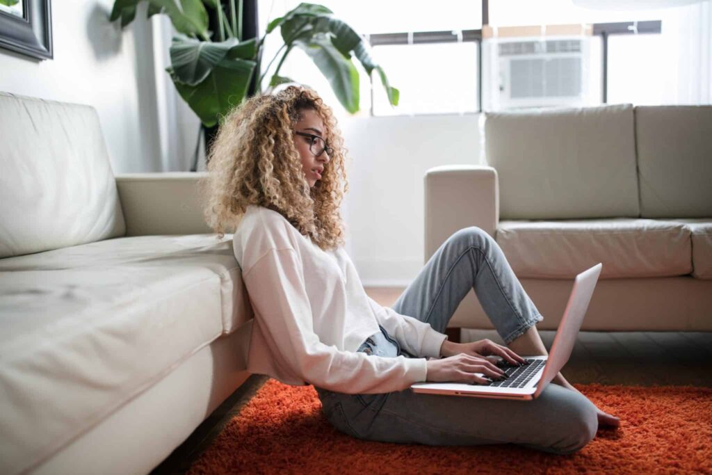 A person with curly hair relaxes on an orange rug, comfortably leaning against a sofa while using a laptop in a bright, inviting living room featuring a large plant and sunny window. Trust Bell Brothers for home comfort—our background-checked technicians proudly serve Northern California communities with upfront pricing and our No Surprise Guarantee.