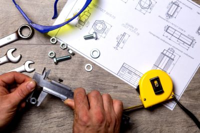 A technician carefully measures a metal bolt on a workbench, surrounded by wrenches, fasteners, safety glasses, and precise technical drawings. This scene reflects Bell Brothers’ trusted service across Northern California—where you’ll always find background-checked technicians, upfront pricing, and our No Surprise Guarantee for complete peace of mind.