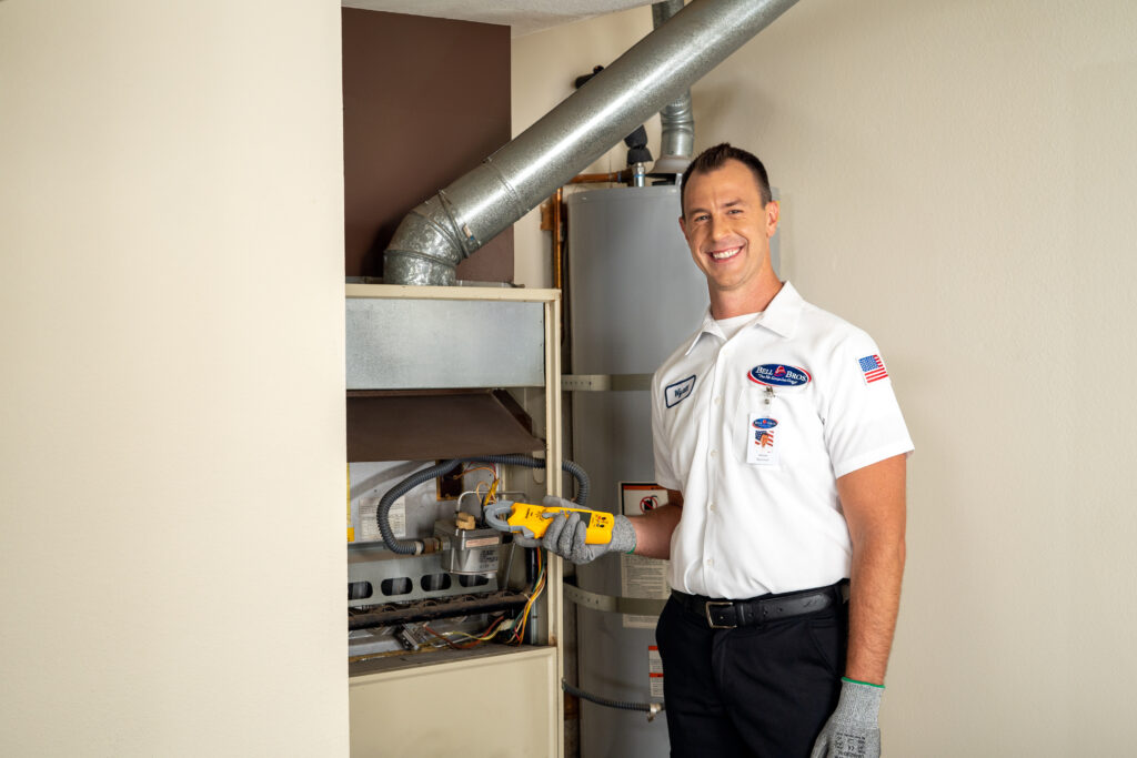 A Bell Brothers technician, dressed in uniform and ready with professional tools, stands beside an open furnace and water heater in a home's utility room—prepared to provide thorough furnace maintenance and ensure your home is cozy for winter. With our trusted No Surprise Guarantee, background-checked technicians, upfront pricing, and outstanding reputation throughout Northern California communities, you can count on Bell Brothers for service you can trust.